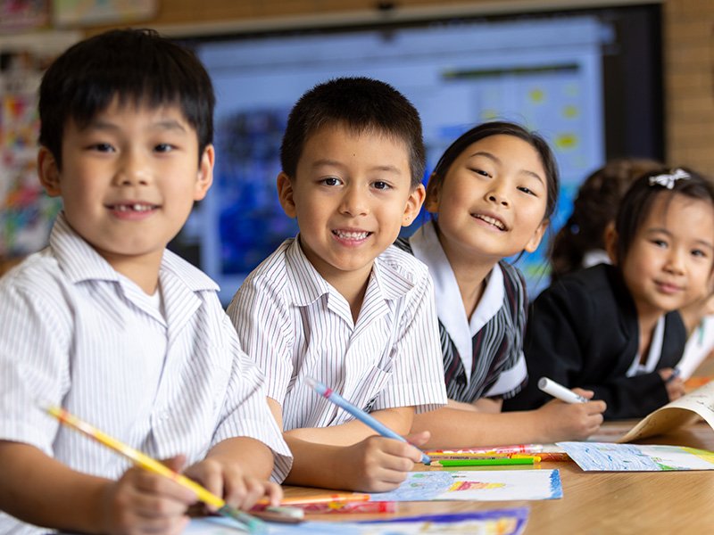 Primary students smiling while sitting at a table in class, colouring in.