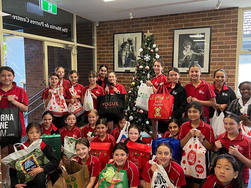 A group of students next to a Christmas tree, holding Christmas gift bags and groceries for charity.