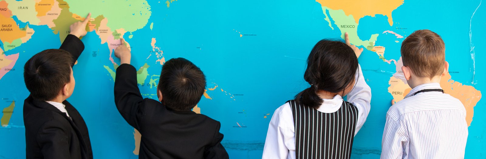 Four primary students facing away, pointing at locations on a world map.