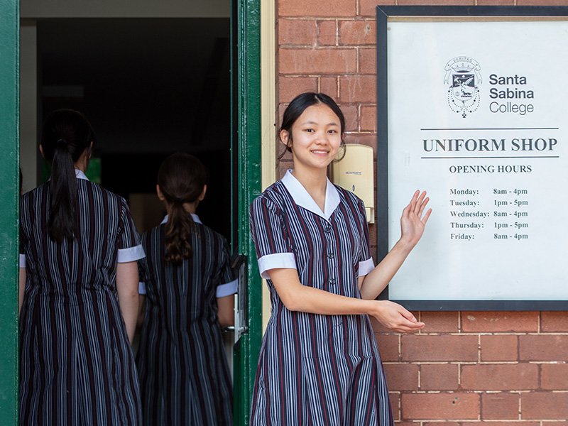 Student standing outside of uniform shop, smiling and pointing at uniform shop sign.