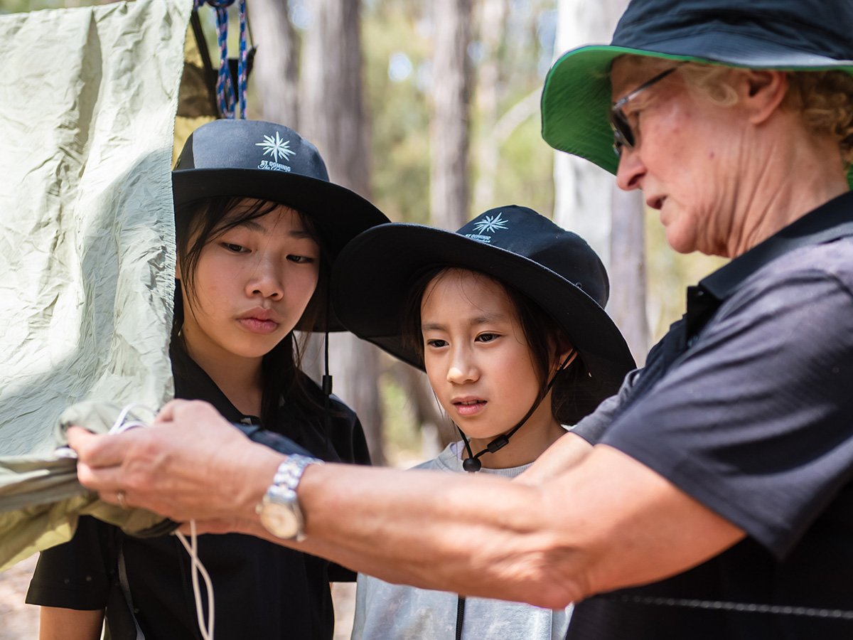 Two primary students watching a teacher demonstrate how to set up a tent.