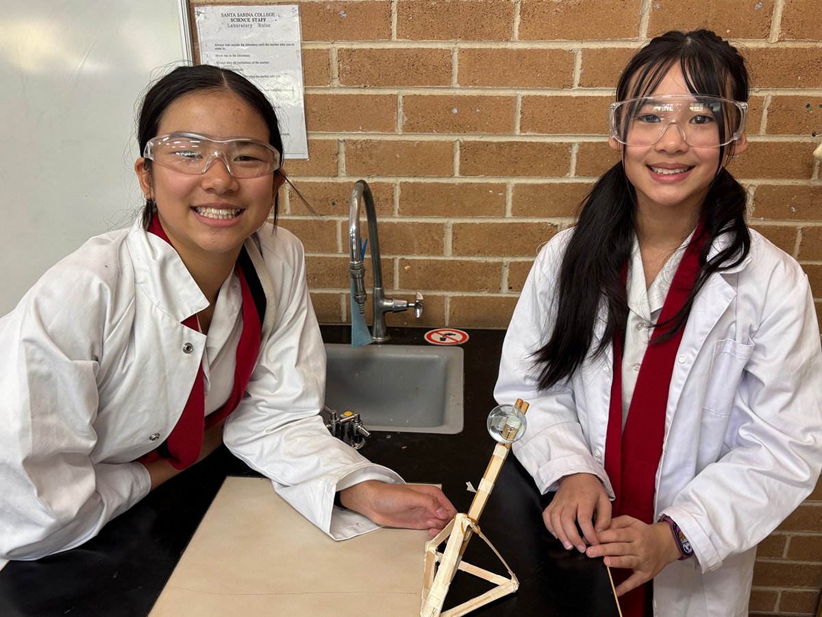 Two students smiling while working on a science project, wearing lab coats and safety goggles.