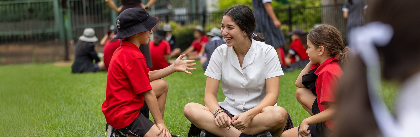 High school student sitting on the grass, smiling and listening to two primary students.