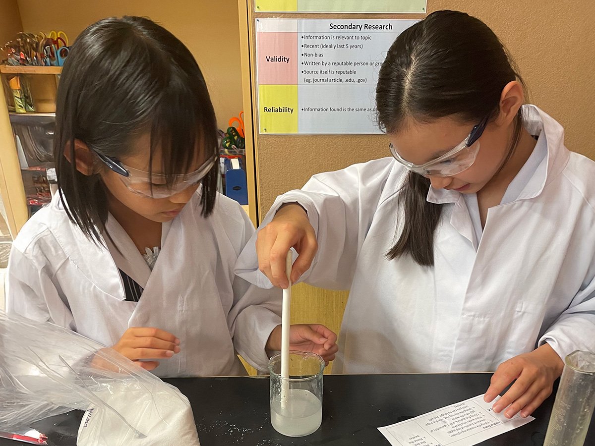 Two students working on a science project, wearing lab coats and safety goggles.