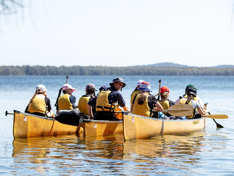 A group of students canoeing on a lake, wearing life jackets.